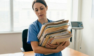 Female nurse stands up, struggling to hold a large pile of insurance denials.