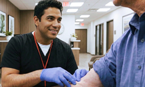 Male phlebotomist kneels down to speak to elderly patient.